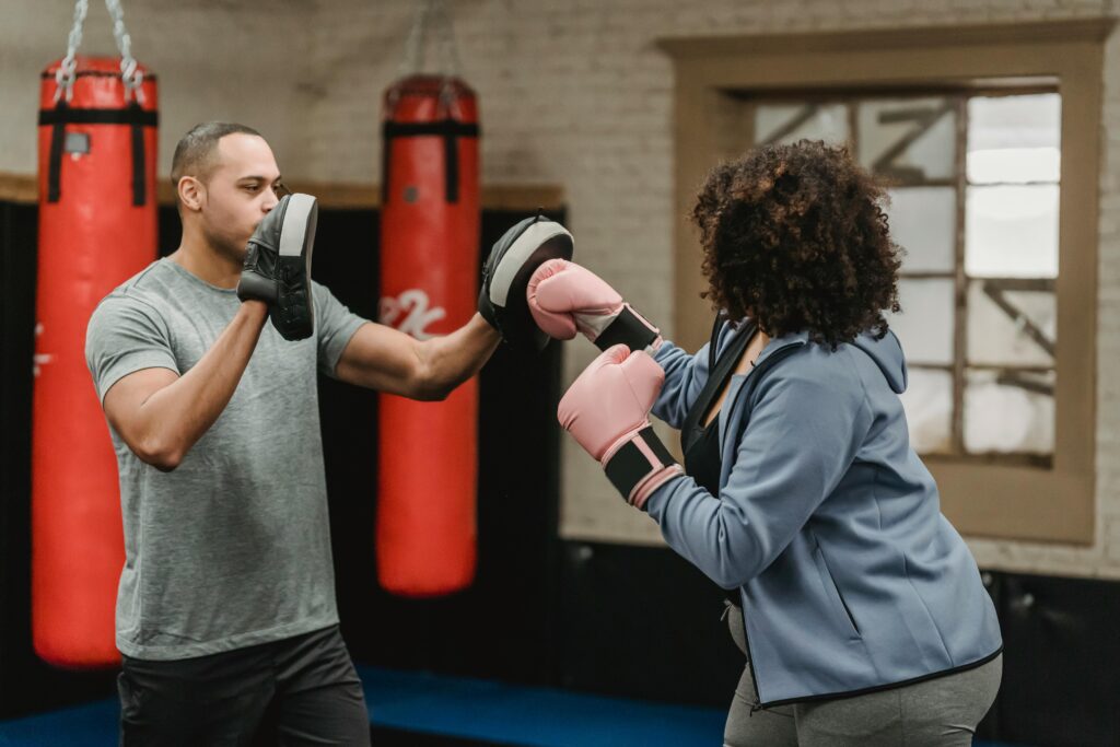Side view of strong African American male personal trainer with boxing paws helping unrecognizable female in gloves during boxing workout
