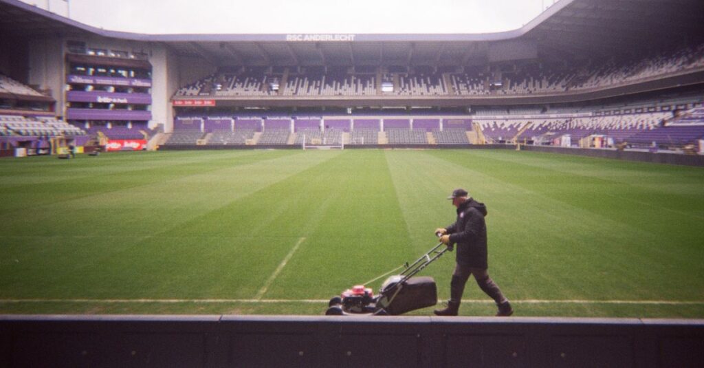 A lone groundskeeper mows a soccer field in RSC Anderlecht stadium, preparing for upcoming sports events.