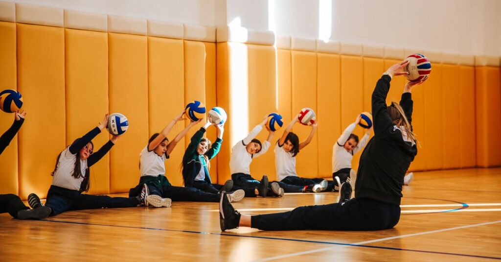 Group of children practicing volleyball stretches in a spacious indoor gym with a coach.
