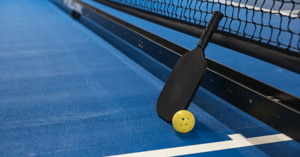 Pickleball paddle resting against net with yellow ball on blue court