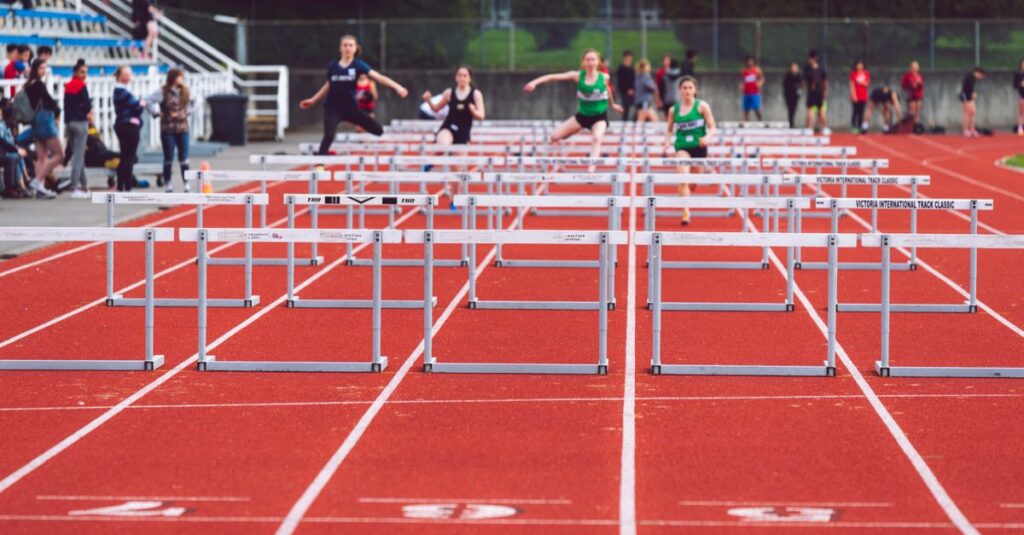 Women athletes compete in a hurdles race at a Victoria track event on a sunny day.