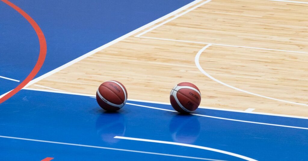 Two basketballs resting on a blue indoor basketball court with wooden flooring.