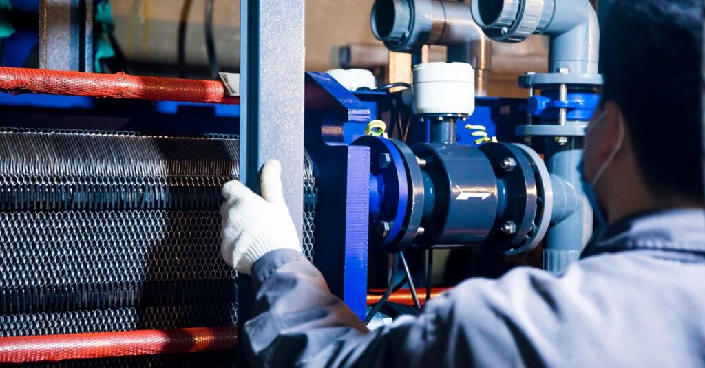 A worker in blue sleeves inspects machinery pipes and equipment at an industrial plant.