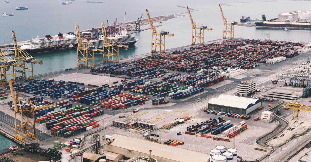 Expansive aerial view of a bustling container port with large cranes and cargo ships under a cloudy sky.