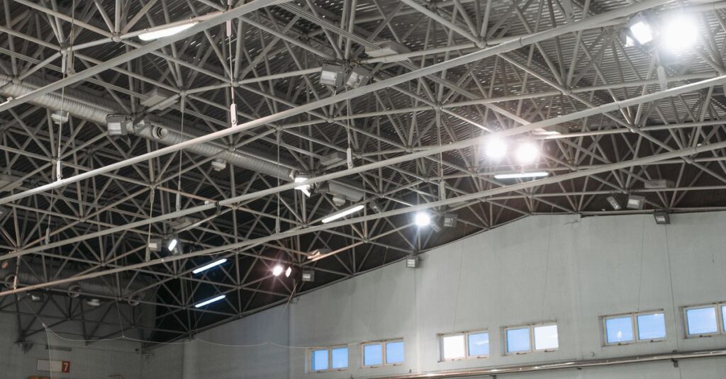 Spacious indoor ice skating rink under a metal truss roof with bright lighting.