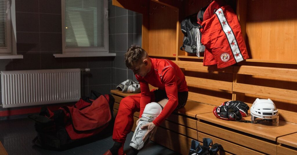 A hockey player in a red uniform prepares for a game in a locker room setting.