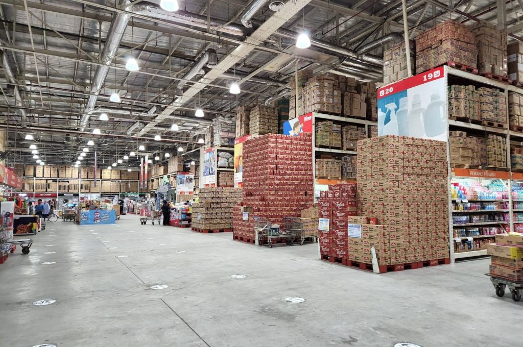 A large wholesale warehouse interior filled with stacked merchandise and shoppers browsing the aisles.