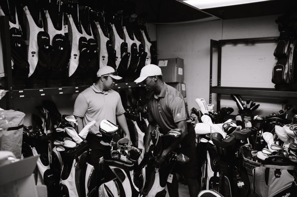 Two men sorting and organizing golf bags and clubs in a storage room.