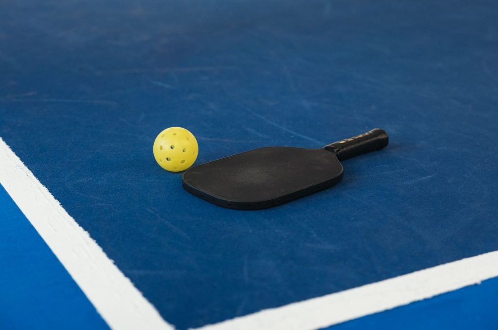 A pickleball paddle and yellow ball on a blue court corner, symbolizing sport and activity.