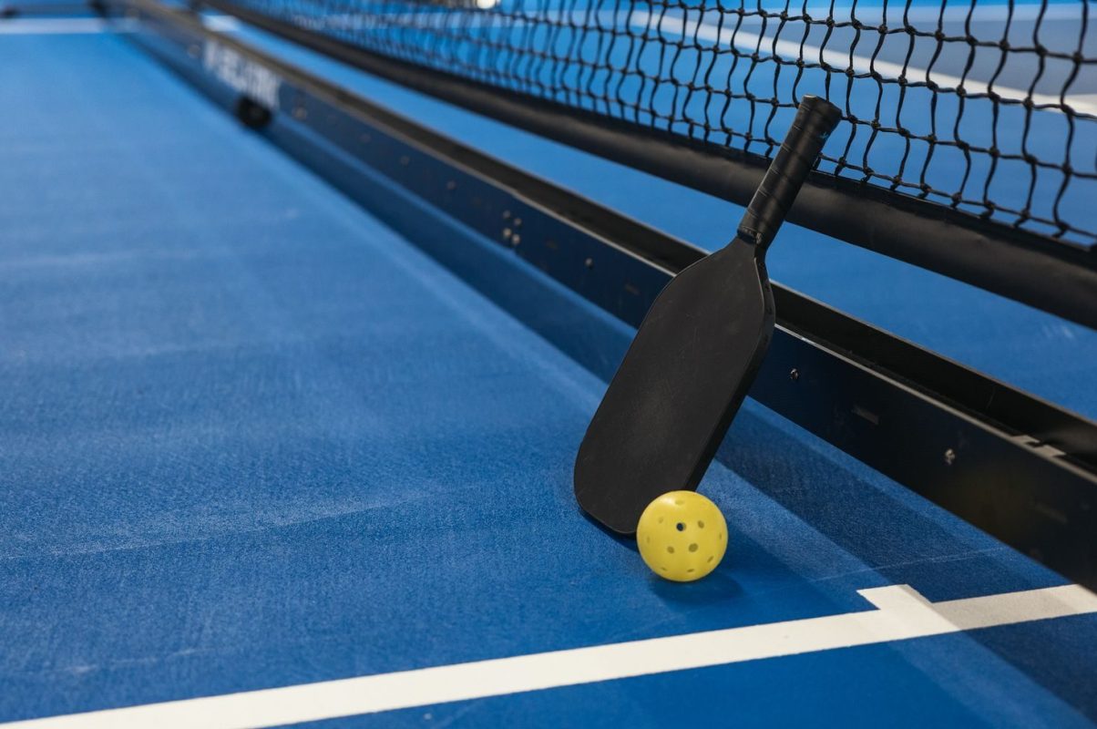 Pickleball paddle resting against net with yellow ball on blue court