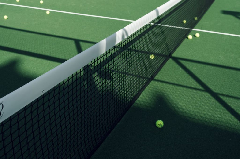 Tennis court net casting shadows on a green surface with scattered tennis balls.