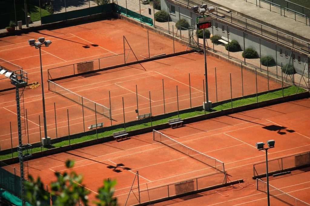 Aerial shot of empty red clay tennis courts on a sunny day with shadows.