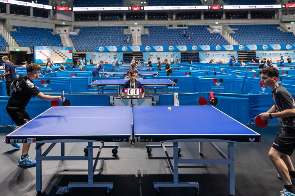 Players compete in a lively table tennis tournament in a large indoor stadium.