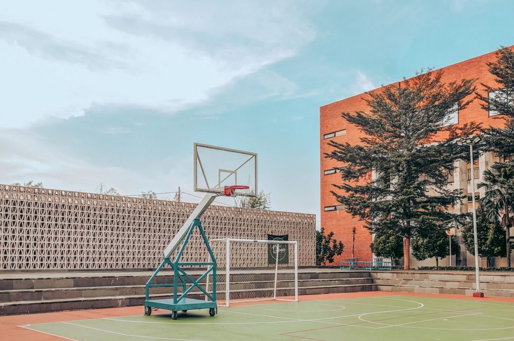 Empty sports ground with basketball hoop and soccer goal on green lawn located near urban building under blue sky