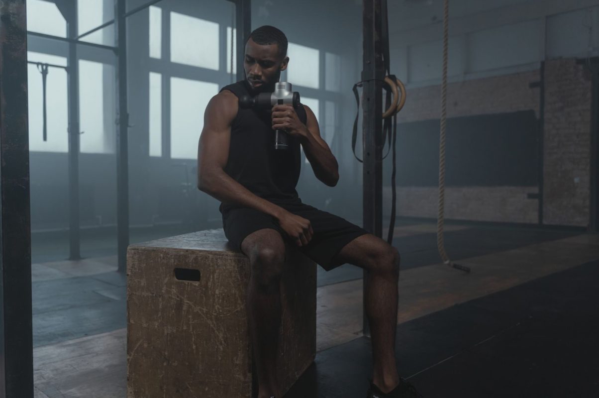 Fit man using a massage gun in a gym, wearing a tank top while seated on a wooden box.