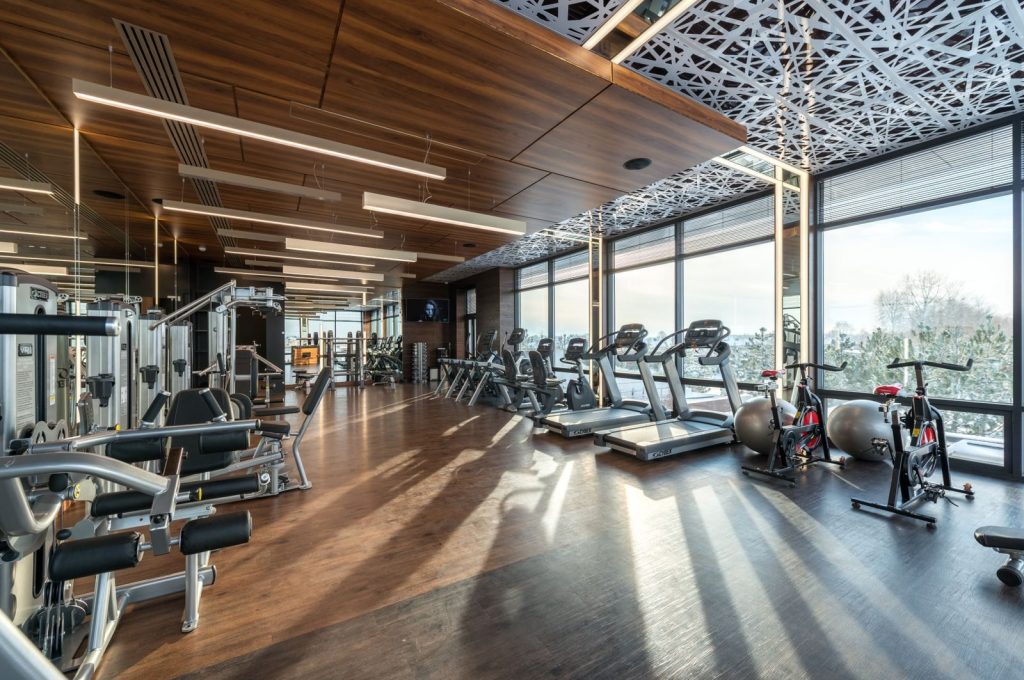Modern exercise bikes and treadmills and various fitness equipment in spacious gym with wooden floor and panoramic windows in sunlight