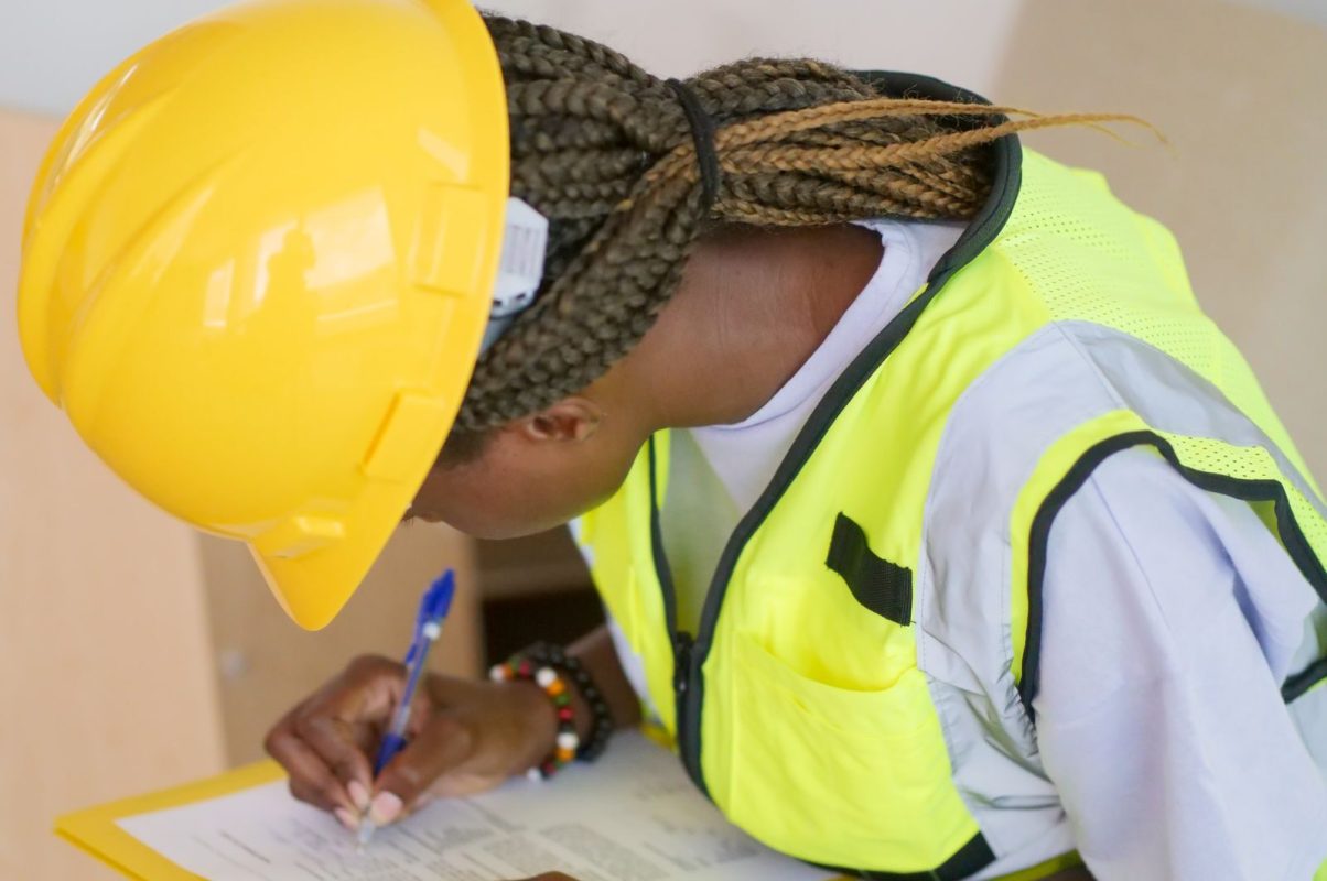 A female engineer in PPE writing on a clipboard at a construction site.